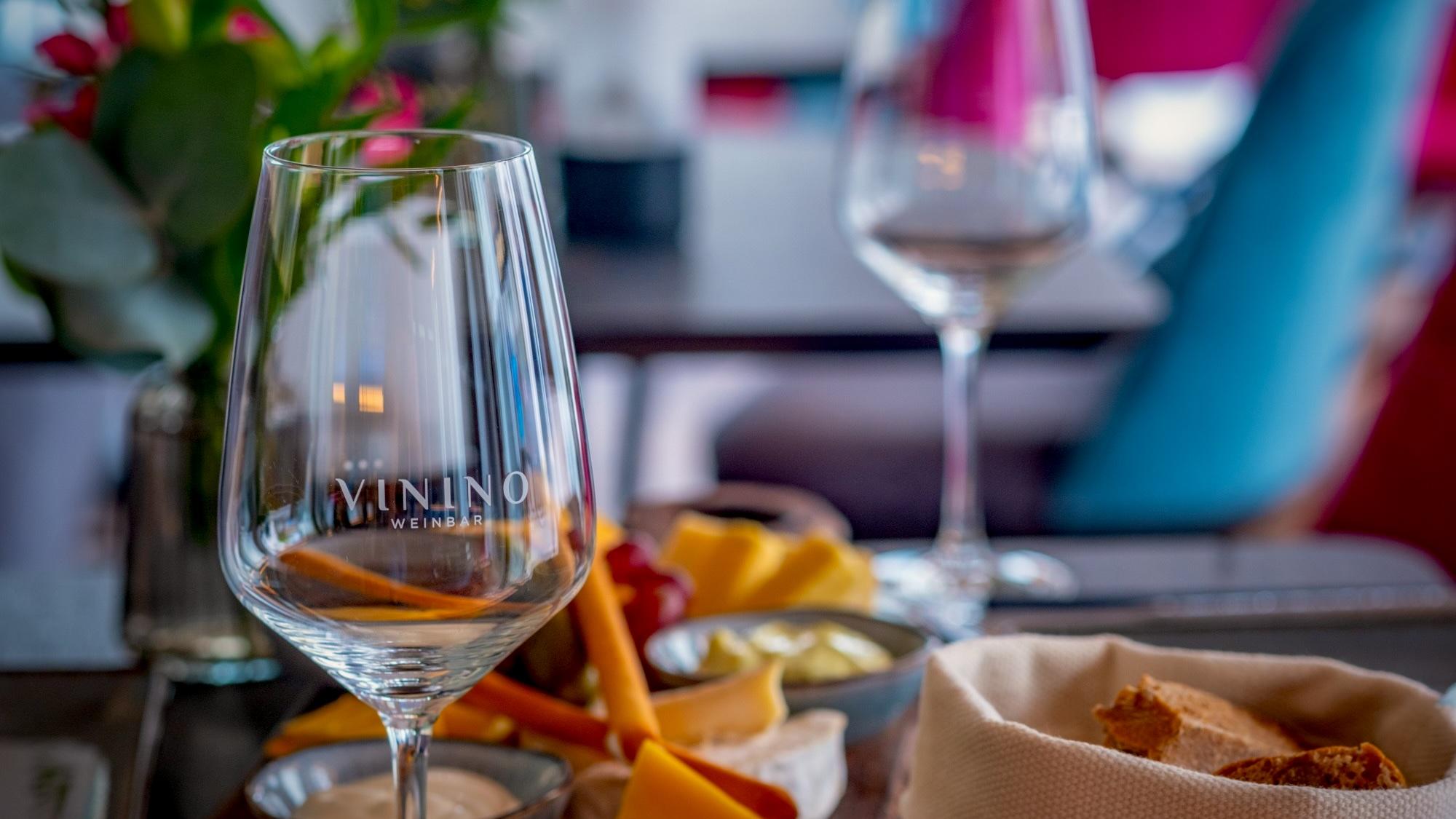 Zwei Weingläser mit VININO-Gravur, Blumen und Käse auf dem Tisch